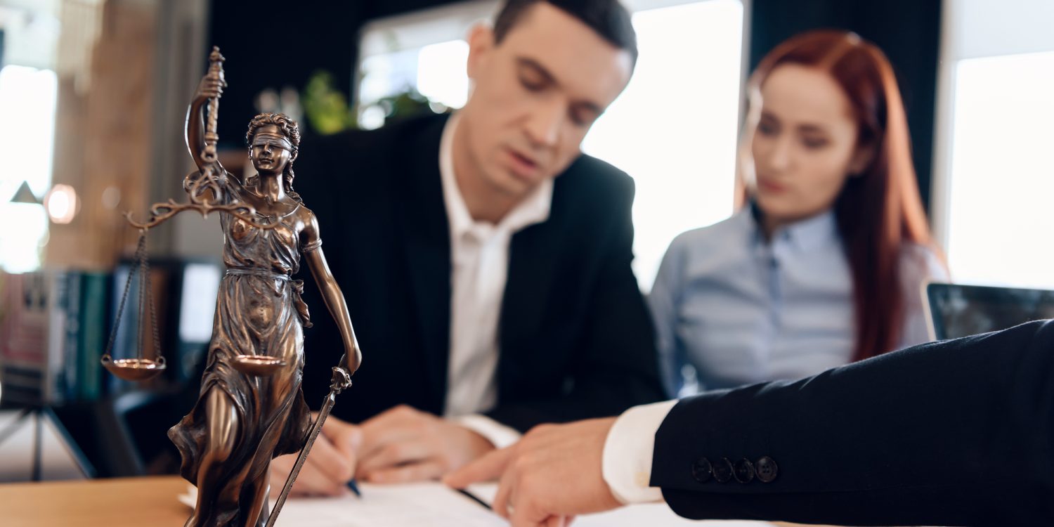 Statue of Themis holds scales of justice. In unfocused background, adult man signs documents. Divorcing couple dissolves marriage contract. Couple going through divorce signing papers.